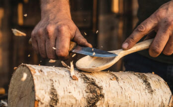 Close-up photograph of an artisan's hands using a sloyd knife to carve a wooden spoon from a raw birch log, with wood chips flying in warm light.