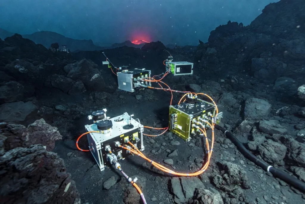 Close-up of deep-sea scientific instruments, including bottom pressure recorders and seismometers connected by cables, on the volcanic seafloor near Axial Seamount.